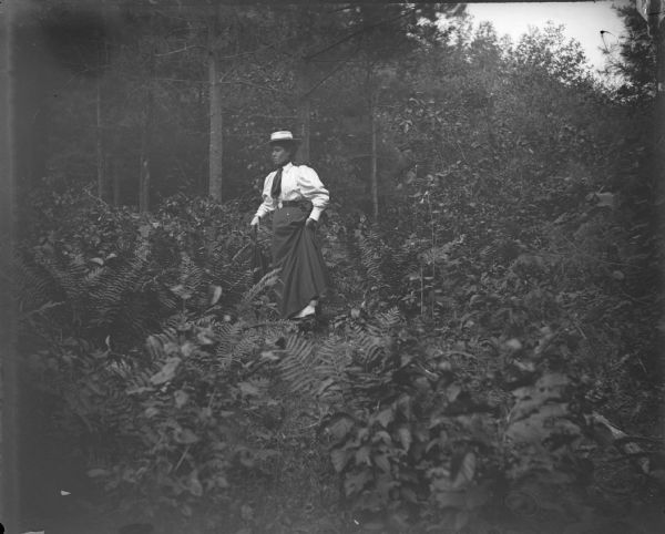 Woman posed standing, holding an umbrella, and lifting her skirt to walk through a field of dense underbrush.