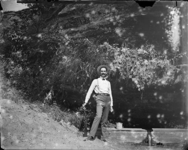 Man posed standing with a gun under an overhanging tree near a small wooden dam on a stream.