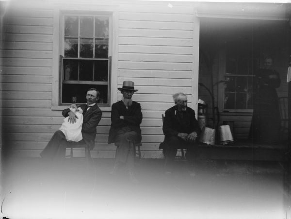 Three men, two elderly, posed sitting in front of a house, one is holding an infant. In the shadows on the right is a woman standing on the porch. Julius Deitrich is holding the baby, Henry Dietrich is center and originally from Prussia, and his father is on the right.