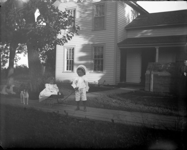 Boy pulling a girl sitting in a wagon followed by a dog on a board sidewalk in front of a house.