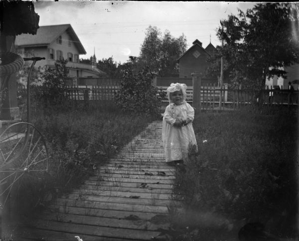 Small girl wearing a bonnet posed standing near a board sidewalk in long grass.