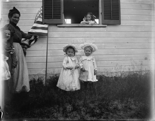 Two small girls holding American flags posed standing on the side of a building, while another girl looks outside from the window. To their side stands a woman with a larger flag and two other children.