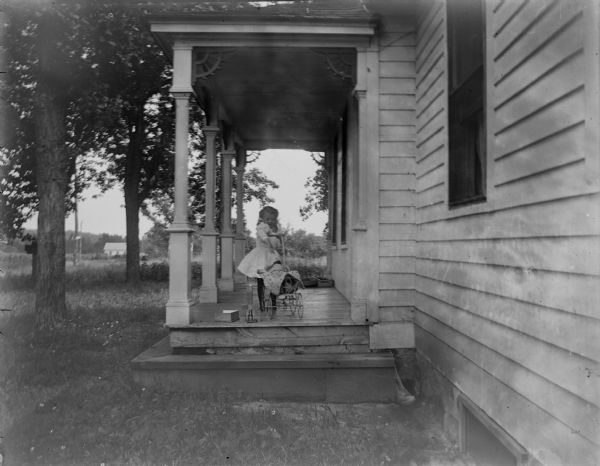 Small girl posed standing on a porch with a doll buggy.