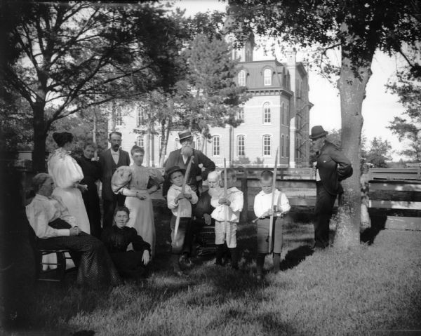 Three boys with archery equipment are surrounded by seated and standing adults in front of the first High School built in 1873.