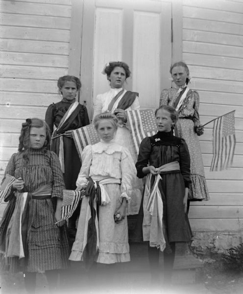 Six girls posed standing and holding United States flags on the steps of a wooden frame building.