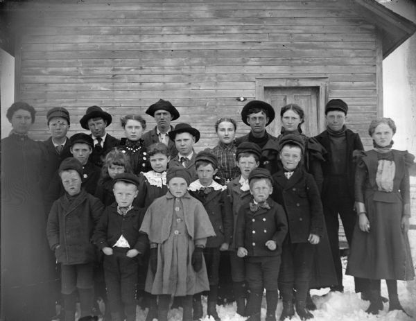 A group of children and a woman, probably a teacher, are posed standing outside of a log building.