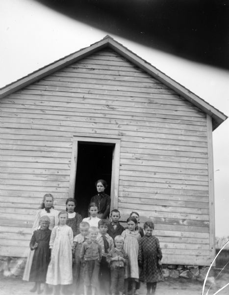 A group of children and a woman, probably a teacher, stand outside of a log building, possibly a schoohouse.