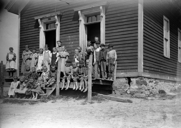 Group of standing and sitting children and a standing woman, probably a teacher, posed on the porch and steps of a building, probably a schoolhouse.
