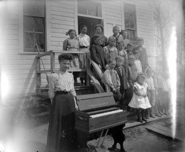 A group of children and woman posed on the steps of a building with an organ, probably a Sunday School class.