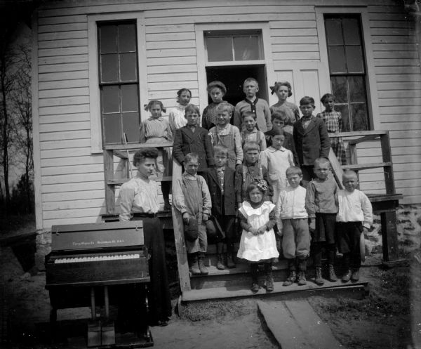 Group of children and woman posed on the steps of a building with an organ, probably Sunday School.