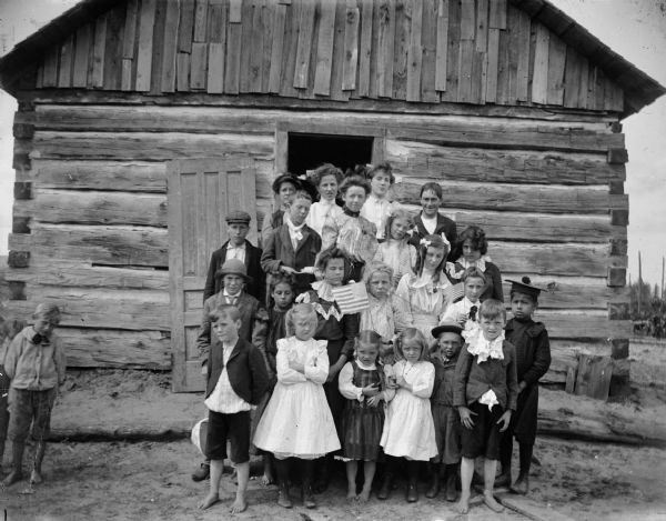 Group of children and a woman, probably a teacher, posed standing on the steps of a log building, possibly in Disco, Wisconsin. One of the children in the center of the group holds an American flag. The teacher is probably Gertie Stratton, who later married a Mr. Hubert.	