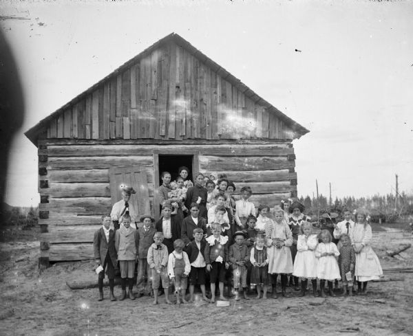 Group portrait of children and a woman, probably a teacher, posed standing on the steps of a log cabin school.
