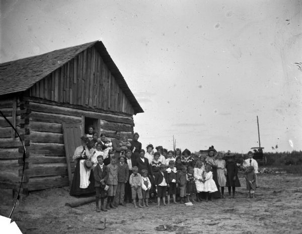 A group of children and a woman, probably a teacher, pose standing on the steps of a log building.