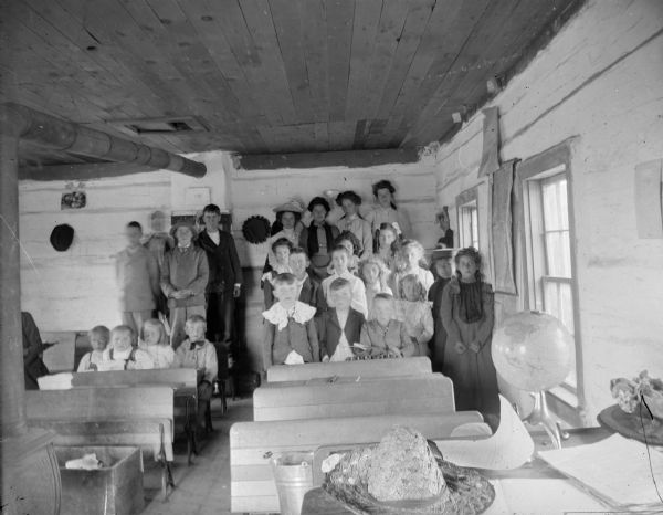 Interior of a schoolhouse, with the children posed sitting and standing in the rear of the building.	