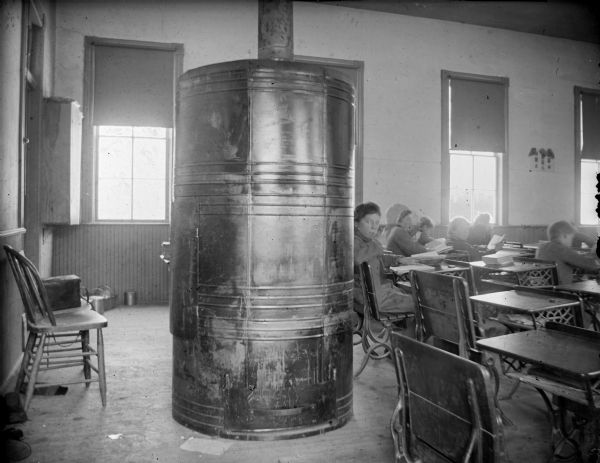 Wood burning furnace at the rear of a schoolhouse classroom. In addition, students are sitting at their desks reading.	