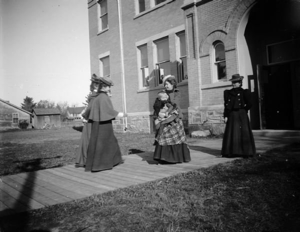 Four women, one holding a young child, stand outside Union High School on a board sidewalk.