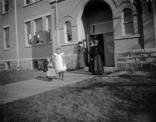 Three women and two girls stand just outside of the Union High School on the boarded sidewalk. Through the windows of the school building, students can be seen in a classroom.