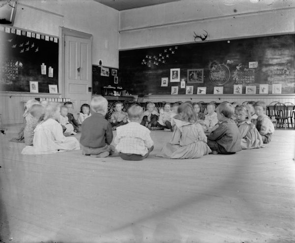 A group of small children, probably Flora LeClaire's kindergarten class, sits in a circle on the floor of a classroom. Various decorations adorn the walls and chalkboards, along with calendars that indicate this photograph was taken in the year 1900.