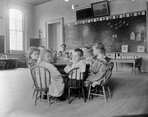 Children  in a kindergarten class sit around a table with their teacher, probably Flora LeClaire. The calendars on the wall indicate that this photograph was taken during the year 1900. 