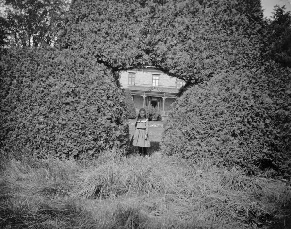 A girl stands in a shrubbery arch in front of a house, probably the yard and house of the Spaulding family.	