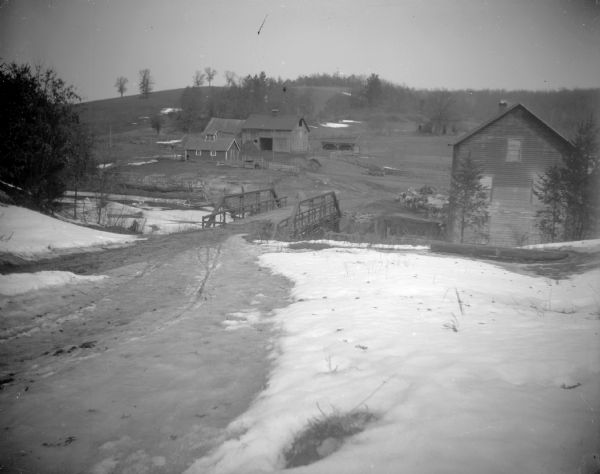 View of a bridge, several wooden frame buildings, and farm land covered with snow. This land is possibly Narracong, later named Loaschings Millon Squire Creek.	