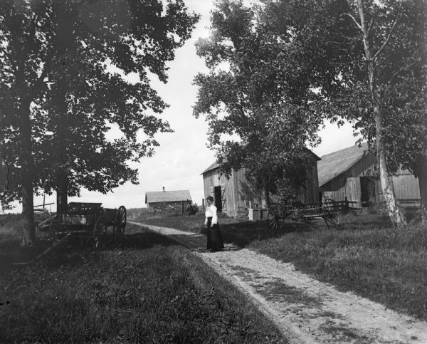 A woman stands on a road that travels through a farm. There are several farm buildings visible along with a wagon.