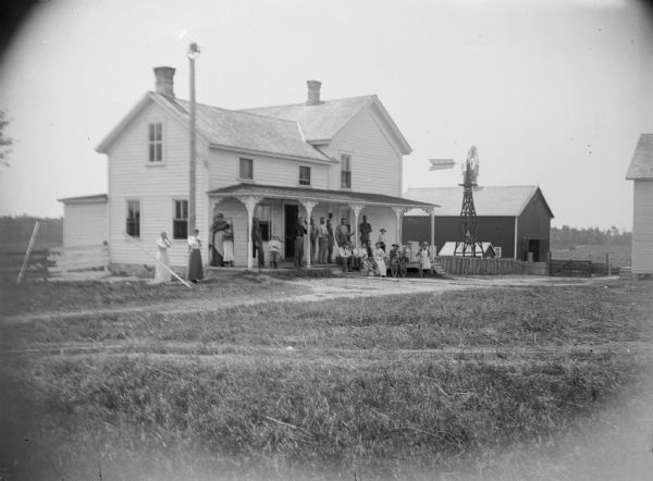 A group of men, women and children gather on the front porch of a farmhouse. In addition to the traditional farm buildings, a windmill is visible near the farmhouse.