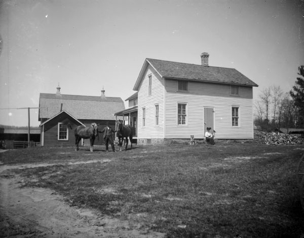 A man poses, displaying two horses in the yard of a simple framed house. A woman sits on the steps of the house with a dog nearby.