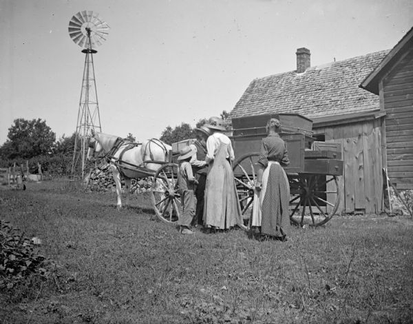 A male peddler shows goods to two women and a young boy. In the background, farm buildings and a windwill are visible.