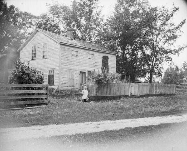 A young girl stands in front of a two-story frame house that is surrounded by a wooden fence.