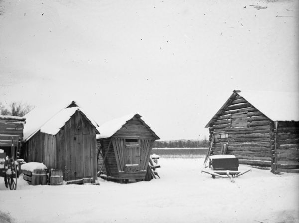 A log cabin is visible on the right, with a corn crib near the center of the photograph. In addition, wooden farm sheds are visible along with various equipment.