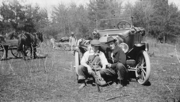 Two men squat in front of an early model Ford inspecting a barren shrub. Two horse-drawn vehicles are seen in the background along with a man and teenage boy.