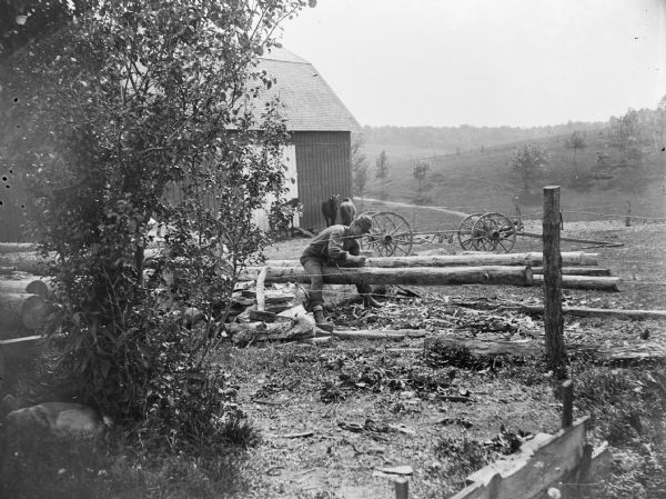 A seated man hews logs in front of a barn. In the background, a rolling landscape is visible.