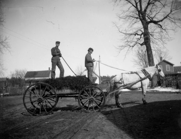 Two men stand atop a wagon of hay that is pulled by a horse. One man holds a pitchfork while the other holds the reins.