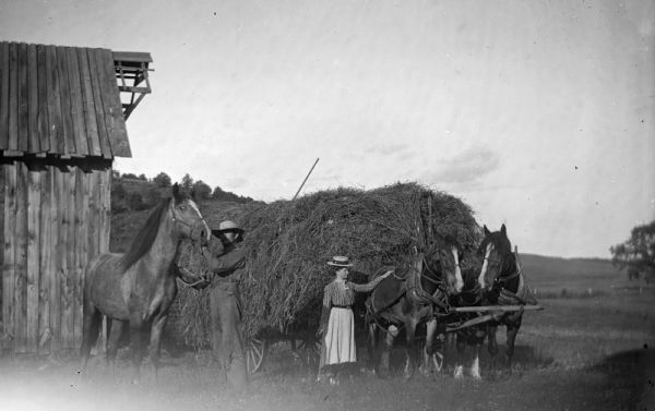 A woman stands with her hand on one of a team of two horses hitched to a wagon overflowing with hay. Additionally, a man stands nearby attending to another horse.