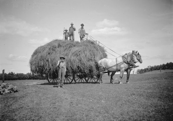 A man stands on top of a hay wagon holding the reins of a two-horse team with a child sitting at his feet. Another man and a boy also pose on the wagon presumably with pitchforks. On the ground, a bearded man stands next to the wagon also holding a pitchfork.