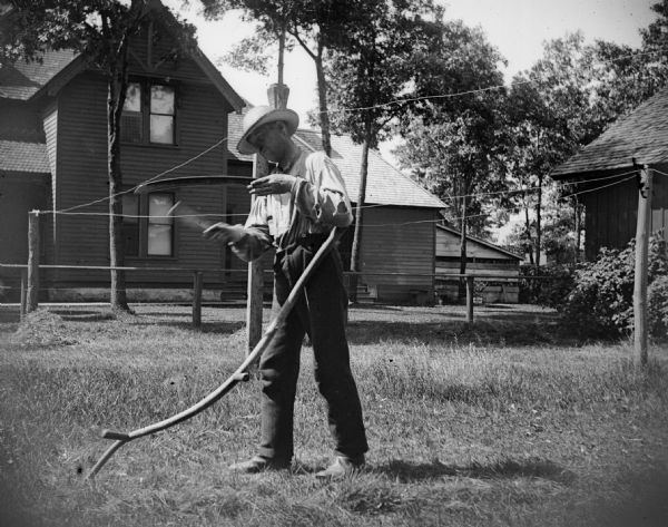 A man using a whetting stone to sharpen a scythe in a backyard.