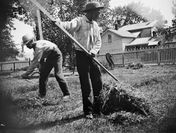 Two boys attend to a lawn, one using a scythe to cut and the other raking the cut grass.
