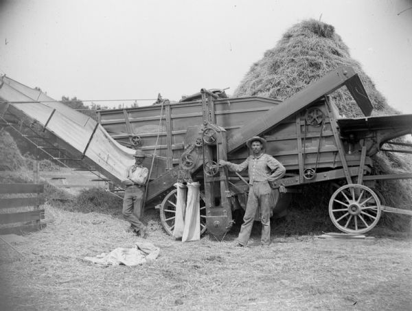 Two men stand near a threshing machine.