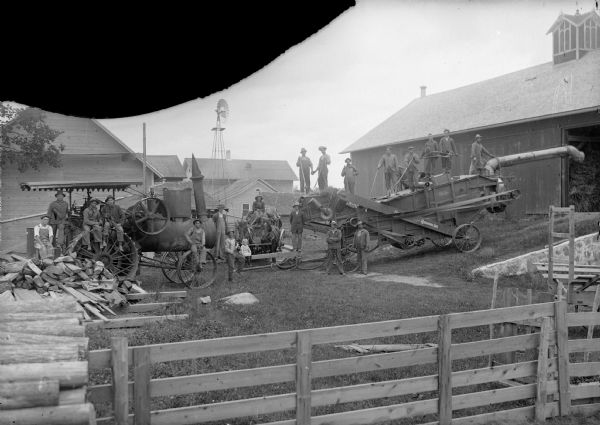 View across fence towards a crew of twenty men posing sitting or standing on a threshing machine and a steam tractor.