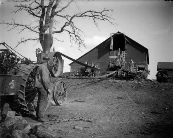 A blower is attached to a steam tractor by a belt and blows grain into a barn. A man stands by the tractor while others stand on the blower near the barn.