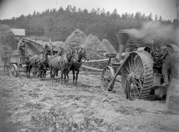 A team of horses working with a steam tractor and threshing machine next to conical stacks of grain.