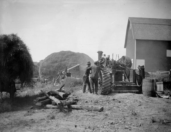 A crew of men inspect a tractor next to a barn, while other men work with a threshing machine in the background.