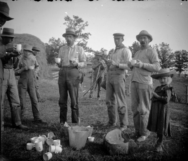 A group of men stand eating and drinking. A little girl also stands with the men while they are on their break from threshing.