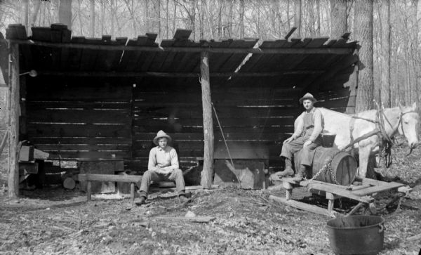 Two men sitting in front of an open-sided wooden shelter. One man is sitting on a barrel on a sled, and the other man is sitting on a log. The sled and horse were probably used to gather maple sap to make maple syrup.