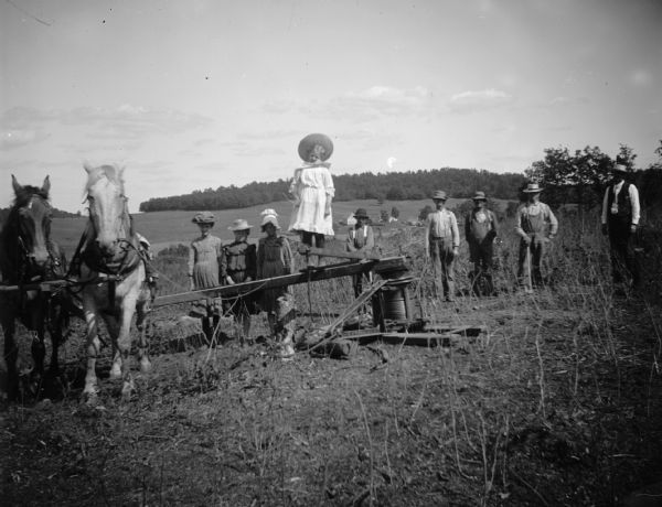 Five men and four girls stand, while one girl poses on a winch turned by a team of two horses. The winch is probably a Svenson Grubber sold by J. H. Carnahan.	