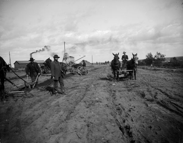 Teams of men and horses work in a field. Two smokestacks and additional agricultural machinery are visible in the background.