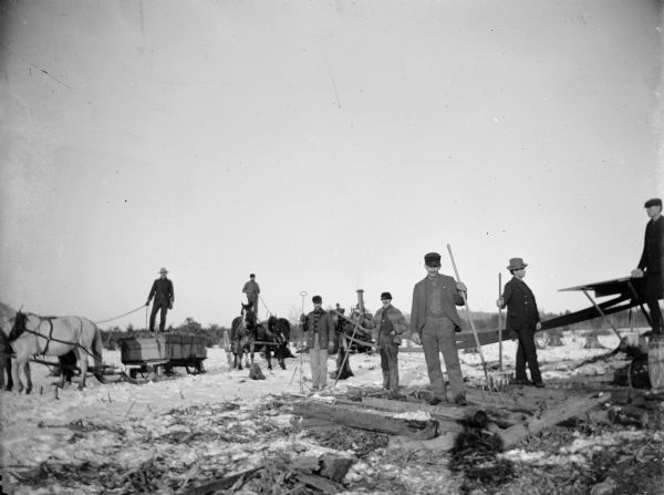 A group of men use steam machinery to clear a field or saw lumber. In the foreground, men stand on a horse-drawn wagon and sled.	