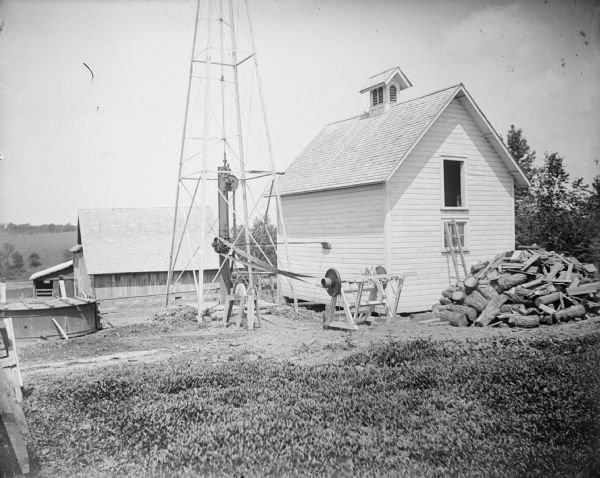 View of a farm with machinery attached to the bottom of a windmill. Windmills were usually used to pump water for livestock. The water was pumped into a tank to cool milk which was kept in very deep cans, 10 inches in diameter and 24 inches deep. From the milk storage tank it flowed into a large wooden tank in the barnyard for the cattle and horses.	