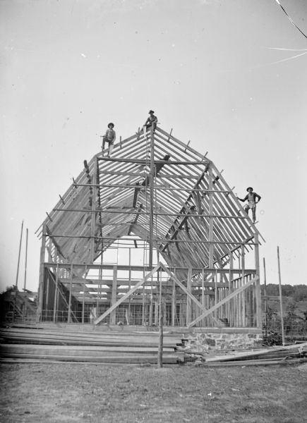 Three men are perched on the framework of a barn.